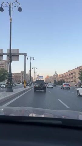 A car POV shot shows a highway with multiple lanes of traffic. The sky is clear and blue, with the sun setting on the right side of the frame. The road is lined with tall, ornate street lamps and large, classical-style buildings with numerous windows. Several cars are driving on the highway, including a black Jeep Wrangler, a white sedan, and a gray sedan. The road appears to be in good condition and the traffic is moderate. The perspective is from inside a vehicle, suggesting the viewer is a passenger.