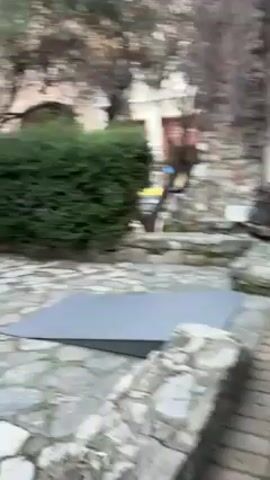 The video pans across an outdoor courtyard with stone paving. In the center is a grassy area with a stone well. The courtyard is surrounded by a covered arcade supported by white pillars with arches above. Trees are visible around the edges of the courtyard.