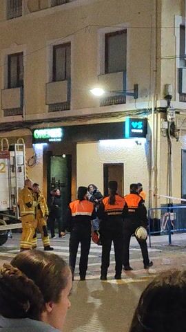 The video depicts an outdoor scene at night, featuring a group of people gathered on a street. In the foreground, there are onlookers, including one with brown hair whose profile is visible. In the street, multiple figures wearing bright orange vests stand around. A fire truck is present, with a firefighter in yellow uniform standing on its platform. The camera is positioned at a low angle, looking up at the scene. Buildings line the street, and streetlights illuminate the area.