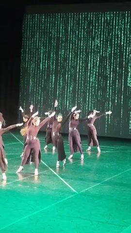 A group of nine women in matching dark purple dance outfits are performing a choreographed routine on a stage. The stage floor is green. In the background, a large screen displays a digital rain effect, reminiscent of the movie The Matrix, with cascading green characters. The dancers are in various poses and movements, some with arms extended, others bending at the waist or kicking their legs. The lighting is predominantly green, casting a glow on the stage and dancers.