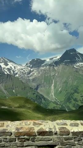 The video shows a scenic mountain landscape. In the foreground, there is a stone wall and some greenery. The middle ground features rolling green hills, some of which still have patches of snow. The background is dominated by majestic mountains, with rocky peaks and snow-capped summits. The sky is blue with scattered white clouds. The camera appears to be moving, possibly from a vehicle, as the perspective shifts slightly. There are no people visible in the video. The environment is clearly an alpine region, likely during the summer or late spring given the presence of both snow and lush green vegetation.