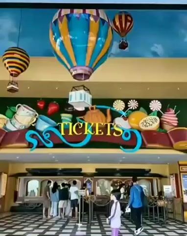 The video shows the entrance to a ticket booth area. The entrance is decorated with large, colorful hot air balloons hanging from the ceiling. Above the ticket windows, there is a large sign that says "TICKETS" in a playful font, surrounded by oversized food items like strawberries, teacups, oranges, and ice cream. The floor is a black and white checkered pattern. Several people are standing in line or walking towards the ticket windows. Some individuals are wearing casual clothing, while others appear to be families. The atmosphere seems to be that of an amusement park or a similar entertainment venue.