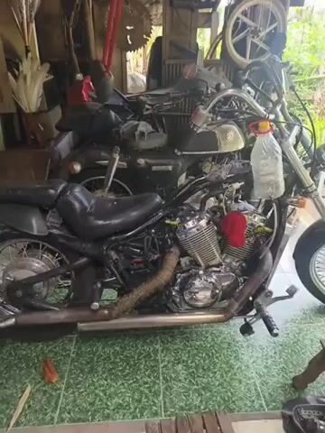The video shows a cluttered garage or workshop containing several motorcycles. The foreground motorcycle is a cruiser-style bike with a black seat and chrome engine details. It has a modified exhaust pipe wrapped in a textured material and a red piece of fabric or plastic tucked into the engine area. A plastic bottle with a red cap is attached to the handlebars. Behind this motorcycle, another motorcycle is partially visible, appearing to be a darker color. The background reveals a variety of items, including tools, mechanical parts like a saw blade, and some foliage outside. The overall impression is one of a working space with projects in progress.