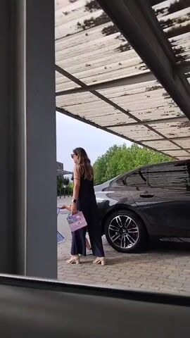 A woman in a dark blue dress stands next to a dark grey car. She is looking to her left. To the right of the car, a man in a red shirt walks away from the camera. Further to the right, a child in a green and blue shirt runs past the woman. The scene is viewed from inside a car, looking out through the driver's side window and side mirror. The area appears to be a covered parking lot with a corrugated metal roof supported by metal beams. Trees and buildings are visible in the background. The ground is paved with interlocking bricks.