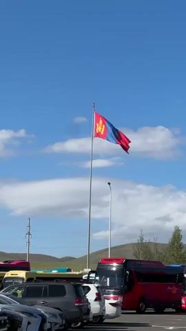 A Mongolian flag flutters in the wind on a flagpole against a clear blue sky with scattered white clouds. In the foreground, several vehicles, including SUVs and buses, are parked in an outdoor lot. The buses appear to be red and white. In the background, rolling hills and trees are visible. The scene suggests an outdoor gathering or travel hub, possibly a rest stop or parking area.