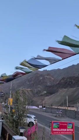 A short video begins with a wide shot of a mountainous landscape under a clear blue sky. Prayer flags in various colors, including blue, red, green, yellow, and white, are strung across the frame, fluttering in the wind. The flags are attached to wires or strings that are supported by poles. In the foreground, a road runs horizontally across the scene, with a few cars parked on the side. A stone wall borders the road. A sign for "HIMALAYAN BITES CAFE & RESTAURANT" is visible. The mountains in the background are rugged and appear to be barren, with slopes covered in loose soil and rocks. The sun is low in the sky, casting a soft light on the peaks. The video pans slightly to the right, showing more of the flags and the mountainous terrain. There are no people clearly visible in the video. The overall impression is one of a scenic, remote location, possibly in the Himalayas.