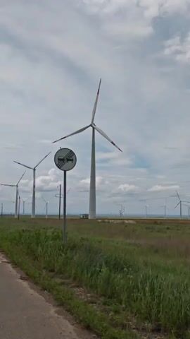 The video shows a line of tall wind turbines in a field, with a paved road running along the edge of the field. The turbines are white with three blades, and some have red tips on the blades. The field is green and grassy, with some taller weeds. Power lines run parallel to the turbines, supported by wooden poles. The sky is overcast with scattered clouds. The camera appears to be moving slowly along the road.