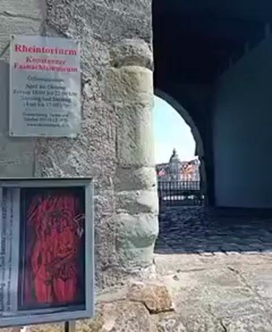A sign with German text is affixed to a stone wall, to the left of an arched doorway. Below the sign is a framed picture of a red artwork. The doorway leads to an area paved with cobblestones, where a distant building is visible. The video is stable and appears to be shot during the daytime.