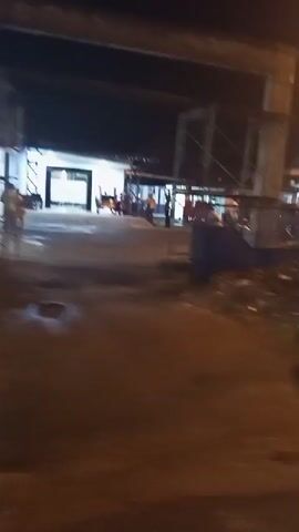 The video shows a blurry night scene from a moving vehicle. In the foreground, there is foliage and darkness. In the middle ground, there are several parked motorcycles and a white sign with text. In the background, there are buildings with lights on and more parked vehicles. There appear to be people standing near some of the motorcycles. The lighting is dim, and the image quality is poor due to motion blur and low light conditions.