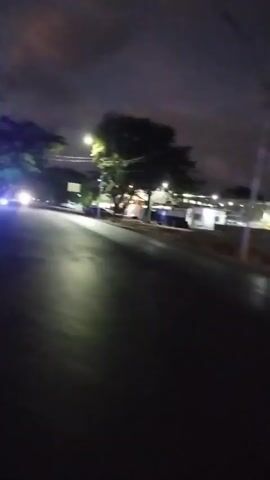 The video shows a nighttime street scene with trees and buildings. There are blue lights illuminating some of the trees and a structure that appears to be a bus stop or shelter. Vehicles with headlights are visible in the distance on the road. The sky is cloudy. The camera is moving, suggesting the video was recorded from a vehicle or while walking.