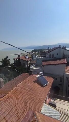 The video shows a view of a coastal town on a sunny day. In the foreground, there are several tiled rooftops of residential buildings. One building has a solar water heater on its roof. Beyond the buildings, a body of water, likely the sea or a large lake, is visible. In the distance, there are hills or mountains. The sky is clear and blue. The camera pans slightly to the right, revealing more of the town and the coastline. There are no people or visible actions in the video.