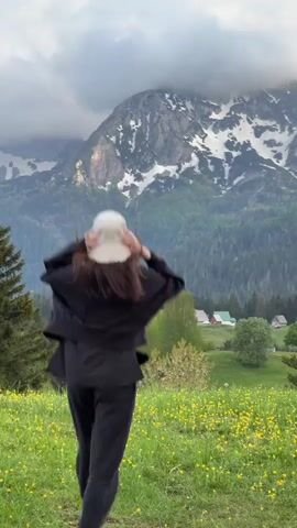 A woman in a black sweatsuit and a white bucket hat runs through a field of yellow flowers. She stops and raises her arms in the air, looking up at a large mountain in the background. The mountain is partially covered in snow and clouds. There are trees in the foreground and a few houses in the distance.