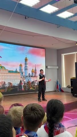 A young boy stands on a wooden stage, speaking into a microphone, with a large LED screen behind him displaying an image of the Moscow Kremlin and its golden domes under a colorful sky. He is wearing glasses, a white shirt with a vest, black pants, and black shoes. He gestures with his hands as he speaks. In front of the stage, the backs of the heads of several children are visible, and some of them are wearing red, white, and blue scarves. To the right of the stage, two Russian flags are propped up. The room appears to be a school auditorium with fluorescent lighting on the ceiling.