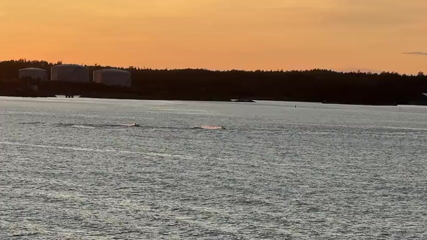 Two jet skis are visible in the water, moving from left to right across the frame. The jet ski on the left is creating a larger wake and kicking up more spray than the jet ski on the right, which appears to be smaller or moving slower. The water is rippled, and the sun is low in the sky, casting an orange glow. In the background, a tree-lined shoreline is visible, with some industrial structures, possibly storage tanks, on the left side. The sky transitions from a pale orange near the horizon to a darker tone further up.