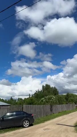 The video shows a rural street scene under a bright blue sky filled with puffy white clouds. A dirt road runs along a weathered wooden fence, behind which is a line of trees and lush greenery. A black car is parked beside the road. Electrical wires stretch across the sky, connected to concrete utility poles. On the other side of the road, a green metal fence borders a brick house with a green roof. Sounds of children playing, including the distinct sound of a ball bouncing, can be heard. The camera pans slowly, showing the landscape and village details.