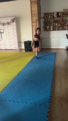 In a dance studio in Sverdlovskiy, Russia, a young girl with long dark hair performs a dance routine. She is wearing a dark crop top and a black fringe skirt, along with dark socks. The studio has a wooden floor and a wooden accent wall. To the left, a large banner with a silhouette of a dancer and the text "DANCING IS LOVE" is visible. In the background, shelves are filled with trophies, suggesting a place of competition or achievement. The girl is moving energetically on a blue mat laid over a yellow mat. The lighting suggests it is daytime. The girl's facial expression is not clearly discernible due to the motion, but her active movements imply focus and engagement in her dance.