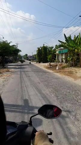 The video shows a first-person view from a motorcycle traveling down a paved road. The road is in a rural or suburban area with houses and vegetation on either side. The sky is blue with some clouds. There are power lines and utility poles visible. In the distance, there are other motorcycles and some people. The motorcycle is moving at a moderate pace, and the camera is relatively stable. The video is short, lasting only a few seconds, and does not show any significant events or interactions.