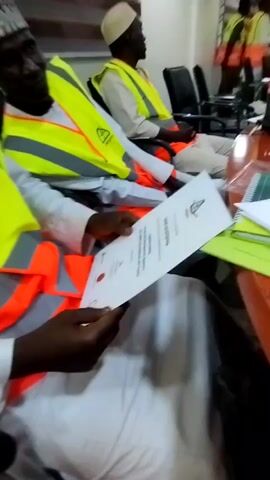 Here is a description of the video:

The video opens with a close-up of a man in a white robe and a bright yellow safety vest, holding a certificate. Another man in similar attire sits beside him. The camera then pans out to show an office setting with several individuals, all wearing similar safety vests. The room is equipped with computers, bottles of water, and other office supplies. Some people are standing and moving around, while others are seated. The overall impression is that of an office environment where a group of people are gathered, possibly for a meeting or training session, as evidenced by the certificate and the formal attire of the participants.