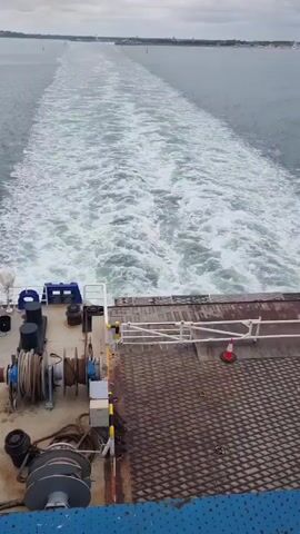 A rear view from a ferry or large boat shows the wake churning behind it in the water. The sky is overcast with gray clouds. The back of the boat has industrial-looking equipment, including large spools of rope or cable and a metal gridded deck. A small orange and white cone is visible on the right side of the deck. The boat is moving away from a distant shoreline with visible buildings.