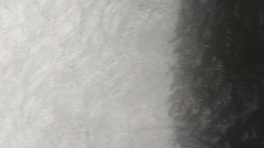 The video provides an extreme close-up, high-angle view of turbulent, white water, likely a waterfall or rapids. The water churns and foams, creating a dynamic and energetic scene. The lighting is dim, with the water appearing bright white in contrast to the darker edges of the frame. There are no discernible people or objects in the water. The environment appears to be natural, with hints of greenery visible at the edges of the frame. The camera angle suggests it is positioned above and looking down into the water.