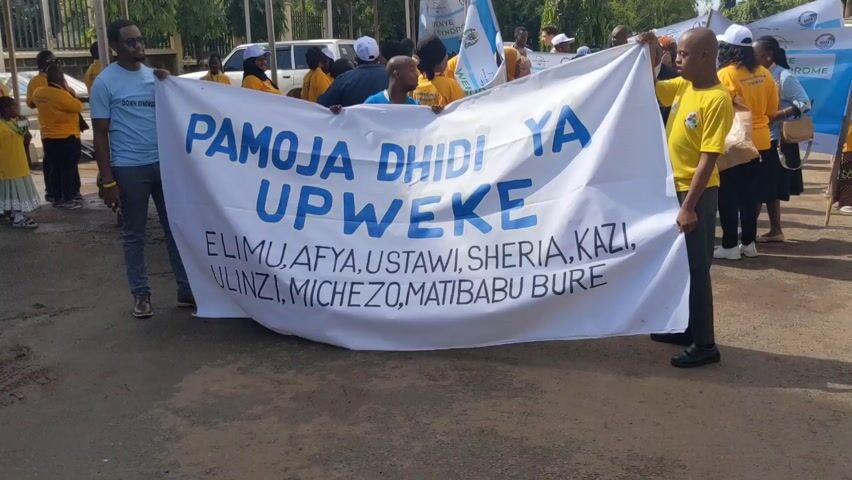The video shows a group of people participating in a march or demonstration.  They are carrying a large white banner with blue text written in what appears to be Swahili. The text reads "PAMOJA DHIDI YA UPWEKE" followed by a list including "ELIMU, AFYA, USTAWI, SHERIA, KAZI, ULINZI, MICHEZO, MATIBABU BURE".  Many participants are wearing yellow shirts. There are also smaller signs with additional text. The event appears to be taking place on a paved road or street, possibly near a building or park. A few vehicles are visible in the background, along with trees and other greenery. The weather looks sunny.