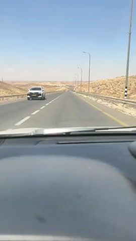 The video shows a view from the inside of a car driving on a highway. The highway has multiple lanes and is bordered by a yellow line on the right and a white line on the left. The surrounding landscape is arid and hilly, with sparse vegetation. There are streetlights lining the side of the road. The sky is clear and blue. The car's dashboard is partially visible in the foreground.