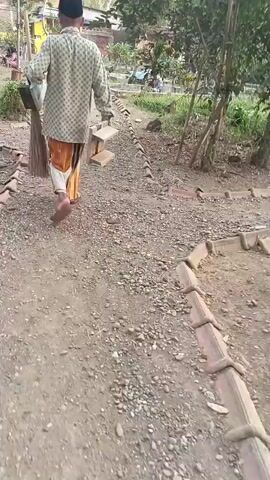 A person is walking away from the camera down a gravel path lined with bricks. The person is wearing a patterned shirt, a sarong, and a black skullcap. They are carrying a broom in their right hand and a brown box under their left arm. The environment appears to be an outdoor area with trees and vegetation.