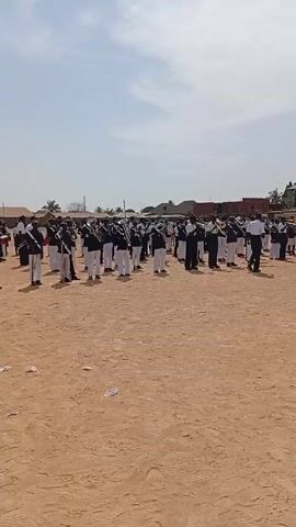 A marching band stands on a dirt field. The members are in black and white uniforms and hold brass instruments. A man in a white shirt walks in front of them. Buildings and trees are visible in the background. The sky is blue with some clouds.
