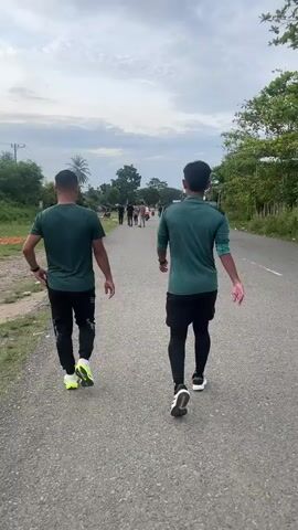 Two men in dark green shirts and black pants walk away from the camera on an asphalt road. The man on the left wears neon yellow and white athletic shoes. The man on the right wears black shorts over black leggings and white athletic shoes. Both men have short dark hair and are walking in the same direction. The road has a white line on the right side. The environment is rural with trees and vegetation on both sides of the road. There are a few other people and motorcycles visible further down the road. The sky is overcast.