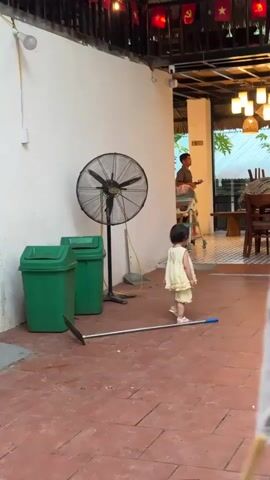 A young child in a light yellow dress and shorts walks away from the camera on a tiled patio. In the background, a man stands near tables and chairs. A large fan and two green trash cans are against a white wall to the left of the frame. A broom lies on the ground in front of the child.