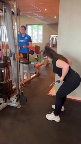 The video shows a woman doing cable rows in a gym. She is bent over, holding the handle of the cable machine, and pulling it towards her abdomen. She is wearing a black top and black leggings, and white sneakers. The gym appears to have standard workout equipment. Behind her, a man stands by another weight machine, appearing to adjust settings. The gym has a neutral interior with a blend of orange, white, and gray color tones.