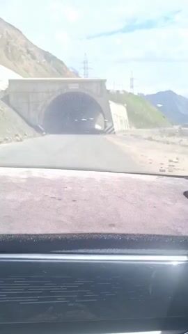 A car drives toward and into a concrete tunnel. The tunnel entrance is arched and has black and white striped barriers on one side. Power lines are visible on a grassy hillside near the tunnel entrance. The tunnel walls are concrete, and it's illuminated by daylight coming from the other end. The dashboard of the car is visible, dusty, and has a dark grey color. Music is playing in the background.