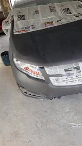 A car is covered in newspaper and plastic sheeting, preparing it for painting. The hood and front bumper are visible, with newspaper masking off the headlights and grille. The surrounding environment appears to be a workshop or garage.