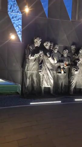 The video showcases a memorial wall adorned with figures holding lanterns, set against a backdrop of geometrically patterned wall and a clear blue sky. The figures, depicted in grayscale, are arranged in a row along the wall, with each figure holding a lantern emitting a warm orange glow. The backdrop features a dark surface with geometric patterns that reflect the sky above. Small lights are fixed above the wall, casting a glow on the figures and the surroundings. The foreground shows a paved walkway leading up to the memorial, with light strips illuminating the base of the wall. The camera slowly pans across the scene, providing a comprehensive view of the memorial and its setting.