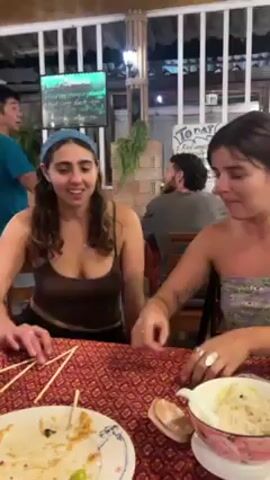 Two young women sit at a table in an outdoor restaurant. The table has a patterned red tablecloth and is littered with used chopsticks and plates of food. One woman is pointing at the other woman's stomach and they are laughing. A waiter in a blue shirt walks behind them. A chalkboard on the wall behind them lists "Today BBQ" with ingredients. There are other patrons sitting at tables in the background. The lighting is dim, suggesting it is evening.