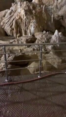 The video shows the inside of a cave with stalactites hanging from the ceiling and stalagmites rising from the ground. A metal railing is visible in the foreground. The cave is dimly lit, and the rock formations are wet and glistening.