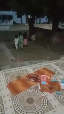 A low-light, grainy video captures a courtyard scene. In the background, several children, possibly four, are visible. One child in a light-colored outfit stands facing left. To their right, another child in a green outfit stands facing right. Next to them, a child in a pink dress also faces right. A fourth child, in a dark outfit, is partially visible, crouched down.

The foreground of the video shows a tiled patio area. Scattered on the tiles are several items: two orange mats, a pile of what appears to be a folded blanket or fabric, and a collection of plastic toys. The toys are brightly colored and include what looks like a pink and blue miniature house or playset, and other smaller toy pieces. The lighting is dim, suggesting it is either dusk or nighttime, and the overall quality of the video is poor. The environment appears to be an outdoor residential area.