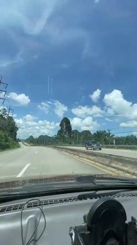 The video shows a view from the inside of a vehicle traveling on a highway. The road is paved and has multiple lanes. The sky is blue with scattered white clouds. On the right side of the road, there are palm trees and a large rock formation in the distance. A few vehicles, including a truck and a car, are visible on the highway. The interior of the vehicle shows the dashboard and windshield.