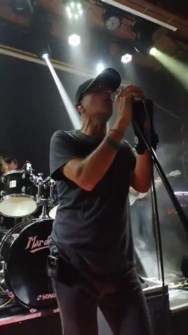 Here is a detailed description of the video:

The video shows a man singing on stage with a band. He is wearing a dark grey t-shirt, dark grey pants, a black baseball cap, and glasses. He's holding a microphone with both hands and appears to be the lead singer of the band. Behind him, a drum set is visible, with the band's name "Mar del S" printed on the bass drum. Other band members can be seen behind him, including a guitarist.

The stage is lit with bright spotlights, creating a smoky atmosphere. The lights change colors, including white, green, and purple. A cameraman moves to show the audience, who are watching the performance with great enthusiasm. Some are holding up their phones to record the show. The camera pans up to show a balcony area filled with people watching the concert from above. The venue appears to be a club or a bar with a wooden interior.