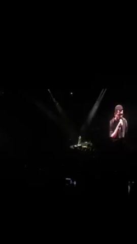 The video captures a live concert from the audience's perspective, dominated by darkness punctuated by spotlights. On stage, a performer stands at a microphone, appearing small in scale relative to the venue's size. A large screen to the right displays a close-up of the performer, revealing him to be a man with dark hair and a beard, dressed in a dark shirt. He holds a microphone to his face, suggesting he is singing or speaking. The crowd is visible as a dark mass of people holding up phones, likely to record the performance. The overall atmosphere indicates a professional, large-scale music event.