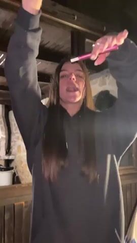 A young woman with long dark hair wears a black hoodie and dances. Her arms are raised and she is looking up and to her right. She is holding a pink object in her right hand, possibly a pen or a wand. The background appears to be an outdoor covered area with wooden beams overhead and what looks like a stone structure to the left. The lighting is dim.