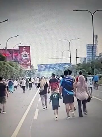 The video shows a group of people walking down a street. The people are of various ages and genders, and they are dressed in casual clothing. The street is lined with trees and buildings, and there are streetlights on both sides of the road. The sky is overcast, and it appears to be daytime. The people are walking in the same direction, and they are all facing away from the camera. Some of the people are walking in pairs or small groups, while others are walking alone. There are also some people in the background who appear to be running or jogging. One person in the foreground is wearing a blue and white shirt and black shorts, and they are carrying a backpack. Another person is wearing a light pink hijab and a light pink top. A young child is walking beside the person in the pink hijab. There are large billboards on the left side of the street, one of which is red and white with an image of a person. In the distance, there are tall buildings. The street appears to be a multi-lane road, and there are white lines marking the lanes.