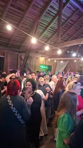 The video shows a crowded indoor event, likely a party or concert, taking place in a rustic wooden structure with a high, beamed ceiling. String lights hang overhead, casting a warm glow, and some of the lights are colored with blue and red hues. The room is filled with people of various ages, many of whom are dancing and socializing. In the background, there appears to be a stage or performance area with lights. Some individuals are holding drinks. The overall atmosphere seems lively and festive.