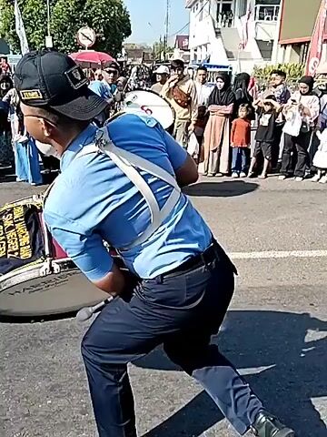 Two men in uniform play large drums and dance in the street, while a crowd of people watches from the side. The first man wears a black cap, light blue short-sleeved shirt, dark pants, and black shoes, carrying a large bass drum with a red and white logo and a blue and gold fabric draped over it. He has a white beard and glasses. The second man wears a light blue short-sleeved shirt, dark pants, and black shoes, carrying a smaller white bass drum with a red and white logo. In the background, there are buildings, trees, street lights, and what appears to be a fountain with water spraying. The street is paved, and the weather is sunny.