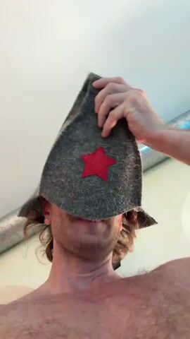 A shirtless man wearing a gray hat with a red star on it is standing in front of an indoor pool. He is looking directly at the camera and making faces, smiling, and flexing his bicep. He has a mustache and light hair. The pool is blue and rectangular. The camera appears to be handheld.