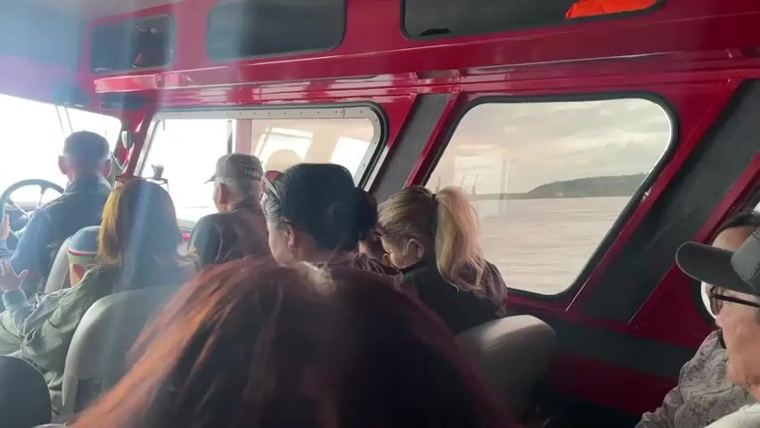 The video shows the interior of a red boat with several people seated. A person wearing a dark shirt is in the driver's seat, holding a steering wheel. The boat appears to be moving, as indicated by the water visible through the windows. The passengers are facing forward, looking out the windows or at the driver. The boat's interior is lined with red panels and has several compartments overhead. The overall scene suggests a boat tour or transportation on a body of water.