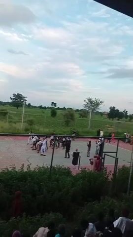 The video shows an outdoor event at dusk. A group of people, some wearing traditional attire, are gathered on a fenced court, possibly for a game or performance. Other spectators are visible in the foreground and on a hill in the background. The sky is cloudy, and floodlights are mounted on poles around the court.