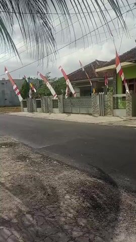 The video shows a street scene with buildings and a large dump truck. There are flags on poles in front of the buildings. The buildings appear to be houses or a small mosque, with green walls and tiled roofs. The dump truck is yellow and green, and it is loaded with what appears to be dirt or gravel. The street is paved with asphalt, and there is a gravel shoulder on the side of the road. The sky is cloudy.