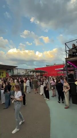 A low-angle vertical video shows a crowd of people gathered outdoors at what appears to be a music event. The sky above is blue with scattered clouds, some illuminated by the setting sun. In the background, a long building with a red facade stretches across the scene, with graffiti visible on it. A large black stage structure dominates the right side of the frame, featuring speaker systems and lighting rigs. A person is visible on the stage behind DJ equipment. A security guard in a white shirt and black pants stands in front of a metal barrier. The crowd consists of many individuals, mostly women, dressed in casual summer attire. Some are standing and facing the stage, while others are milling about. A woman in the foreground, wearing a white t-shirt and blue jeans, is facing away from the camera and to the left. The ground is paved and appears to be an outdoor open area. The overall atmosphere suggests a lively evening event.