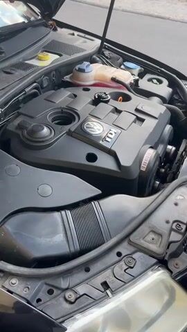 The video shows a close-up, overhead view of a car engine bay with the hood open. A person's hands are visible as they perform a car maintenance task, likely an oil change. The engine appears to be a Volkswagen TDI, indicated by the "TDI" badge on the engine cover. A bottle of engine oil, labeled "MAXOIL 5W-30," is being held and poured into the engine. The scene suggests a daytime setting, possibly a driveway or a workshop, given the natural lighting and the street visible in the background. The focus is on the mechanical aspect of the activity, with no discernible emotional cues from the person or any specific weather conditions. The engine compartment includes various components such as the oil cap, coolant reservoir, and hoses, all characteristic of a modern internal combustion engine. The overall impression is one of routine automotive upkeep.